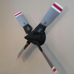 Pode incluir: Um suporte de parede com hélice de avião de quatro pás, apresentando um cubo central preto fosco e pás cinzentas. Cada pá tem uma ponta com listras vermelhas e brancas, um pequeno emblema circular perto da base e texto técnico impresso.