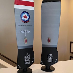 May include: Two vertical aircraft propeller blades mounted on black circular stands. The blades are grey with black bases, featuring red and white stripes at the top, military-style circular emblems, wing insignias, and technical text including NO STEP.
