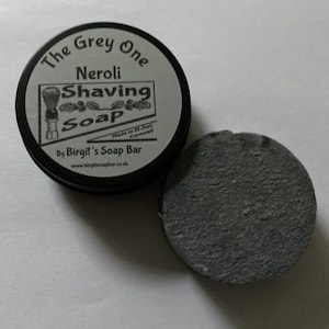 May include: A round, gray shaving soap puck next to a black tin. The tin lid reads "The Grey One Neroli Shaving Soap" with a graphic of a shaving brush. The soap is made by Birgil's Soap Bar.