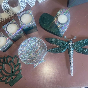 May include: A collection of decorative items, including tealight candle holders, a heart-shaped candle holder, a seashell-shaped dish, a dragonfly figurine, and a lotus flower-shaped coaster. The items are decorated with glitter and are on a brown surface.