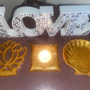 Puede incluir: Una composición decorativa con un letrero "LOVE" blanco adornado con pedrería. Acompañando el letrero hay una flor de loto dorada, una vela en un soporte dorado en forma de almohada y un cuenco dorado en forma de concha.