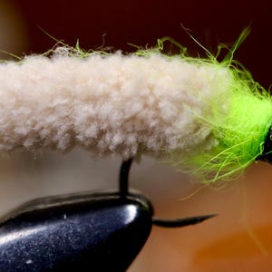 May include: Close-up of a fly fishing lure. The lure has a cream-colored, fluffy body and a bright green head. A black hook extends from the lure's body. The fly is designed to mimic an insect.