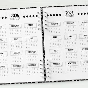 May include: Open notebook with 2026 and 2027 calendars. Each month is displayed in a box. A pencil and a binder clip are on the right side. The notebook has a black and white patterned border.