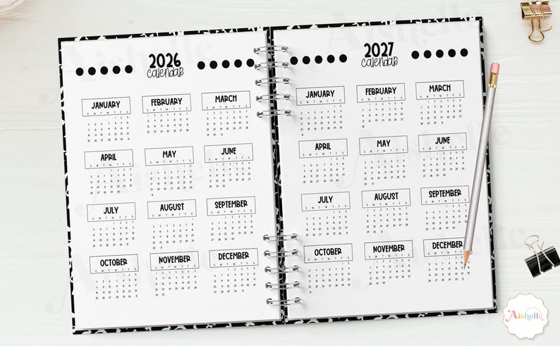 May include: Open notebook with 2026 and 2027 calendars. Each month is displayed in a box. A pencil and a binder clip are on the right side. The notebook has a black and white patterned border.