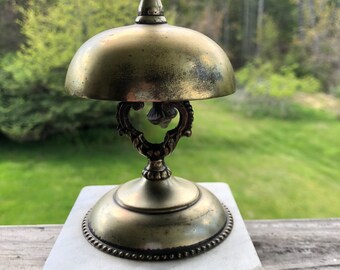 May include: Antique brass bell with a domed top, ornate handle, and a white marble base. The bell has a tarnished finish, suggesting age. The background is a blurred view of green trees.