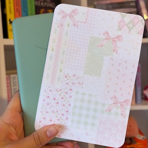 May include: A rectangular e-reader device with a solid sage green back casing, partially covered by a decorative rectangular card featuring a collage of pastel pink and light green patterns, including gingham, floral prints, and three pink ribbon bows.