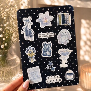 May include: A black sticker sheet with white polka dots featuring various illustrations including ballet slippers, a lily, a row of books, a bunny, tulips, a gummy bear, a cocktail glass, stars, a candle, and text reading so many books, so little time.