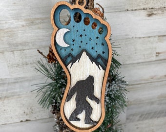 Könnte beinhalten: Weihnachtsornament aus Holz mit Bigfoot-Motiv. Der Ornament hat die Form eines Fußabdrucks, mit einer Silhouette von Bigfoot, Bergen, einem Mond und Sternen. Der Ornament besteht aus geschichtetem Holz mit natürlicher Holzmaserung.