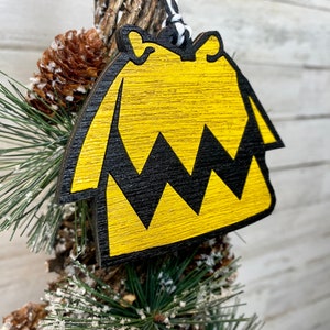 May include: Wooden ornament featuring a yellow and black design resembling a cartoon character's shirt. The ornament is hanging with a black and white string, and is surrounded by pine needles and a pine cone.