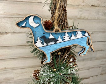 Puede incluir: Adorno de madera con forma de perro salchicha con cuerpo azul y escena nocturna. El adorno presenta una luna creciente, estrellas, un pájaro y un bosque. Cuelga de una cuerda.