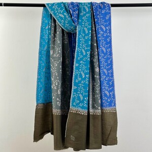 May include: A multi-colored scarf with a floral pattern. The scarf features shades of blue and gray with a brown border. The floral design is white. The scarf is hanging from a black rod.