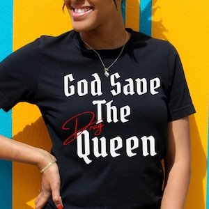 Puede incluir: Una camiseta negra con el texto "God Save The Drag Queen" en letras blancas. La palabra "Drag" está escrita en cursiva roja.