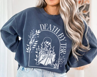 May include: A dark blue sweatshirt with a white graphic of a skeleton reading a book. The graphic is surrounded by leaves and the text "Death by TBR".