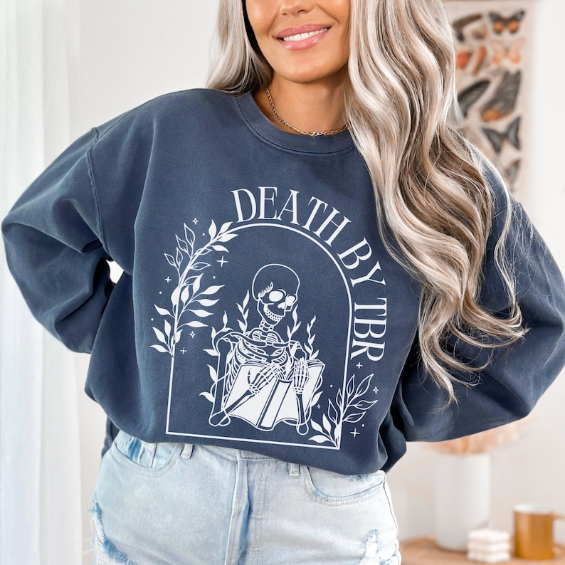 May include: A dark blue sweatshirt with a white graphic of a skeleton reading a book. The graphic is surrounded by leaves and the text "Death by TBR".