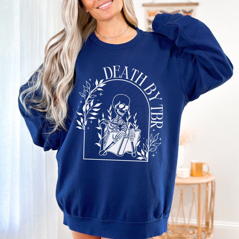 May include: Navy blue sweatshirt with a white graphic of a skeleton reading a book with the text "Death by TBR" above the skeleton.