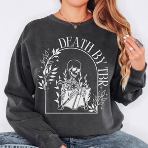May include: A black sweatshirt with a white graphic of a skeleton reading a book. The graphic is surrounded by leaves and stars. The text "DEATH BY TBR" is written in a white arch above the skeleton.