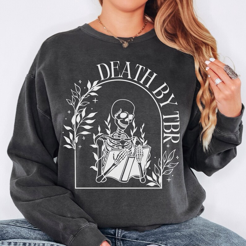 May include: A black sweatshirt with a white graphic of a skeleton reading a book. The graphic is surrounded by leaves and stars. The text "DEATH BY TBR" is written in a white arch above the skeleton.