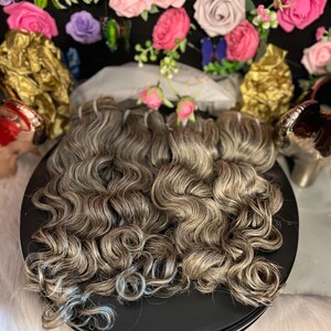 Op de afbeelding: Een bundel golvend echt haar extensions in lichtbruin en grijs. Het haar is in een cirkelvormige patroon op een zwart oppervlak gerangschikt.