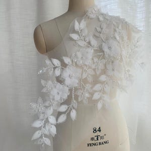 Op de afbeelding: Witte bloemen kanten applicatie met 3D bloemen, bladeren en kralen accenten. Het delicate ontwerp wordt getoond op een mannequin, waarbij de details en textuur worden benadrukt. Ideaal voor bruidskleding of speciale gelegenheden.