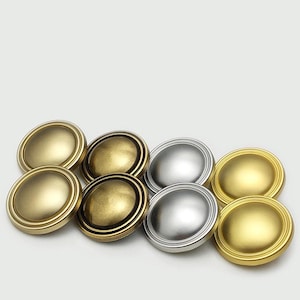 Op de afbeelding: Een verzameling van acht ronde knopen gerangschikt in twee rijen. Vier knopen hebben een matte gouden afwerking, twee hebben een geborsteld antiek messing uiterlijk met donkere randen en twee tonen een glad, zilverkleurig metalen oppervlak.