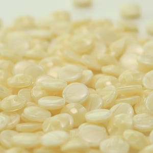 Könnte beinhalten: Nahaufnahme eines Haufens hellgelber Wachsperlen. Die Perlen sind klein, rund und haben ein leicht durchscheinendes Aussehen. Das Bild ist gut beleuchtet und hebt die Textur und Form der Wachsperlen hervor. Die Perlen sind verstreut.