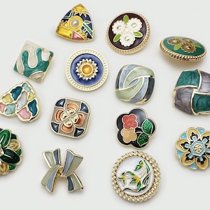 Może przedstawiać: Kolekcja ozdobnych guzików o różnych kształtach i wzorach. Guziki mają złote metalowe obramowania i kolorowe emaliowane inkrustacje. Wzory obejmują wzory kwiatowe, geometryczne i abstrakcyjne, z różnorodnością kolorów.