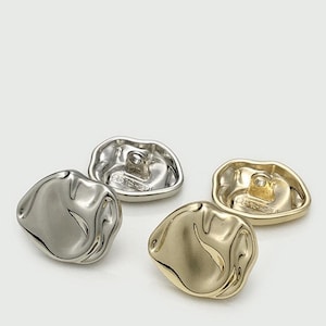 Puede incluir: Cuatro botones metálicos con forma orgánica e irregular. Dos son plateados y dos dorados. Los botones tienen una superficie texturizada y ondulada y un lazo en la parte posterior para coser. Los botones son probablemente para ropa o proyectos de manualidades.