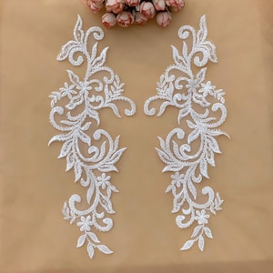 Puede incluir: Dos apliques de encaje floral blanco con intrincados diseños de patrones arremolinados, hojas y pequeñas flores. Los apliques están adornados con pequeños detalles brillantes. Un grupo de flores rosas y blancas es visible en el fondo.