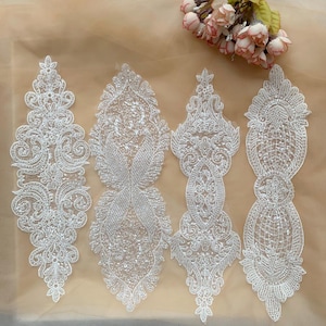 Puede incluir: Cuatro apliques de encaje blanco con intrincados diseños florales y geométricos. Cada aplique presenta un detallado trabajo de cuentas y lentejuelas, creando un efecto brillante. Los apliques están dispuestos sobre un fondo neutro, con un pequeño arreglo floral.
