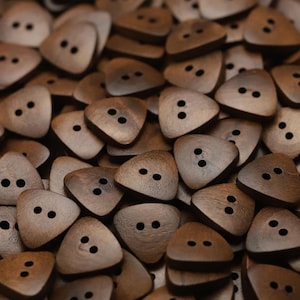 Pode incluir: Uma coleção de botões triangulares de madeira com dois orifícios pretos cada. Os botões são de cor castanho claro com grão castanho mais escuro. Os botões estão espalhados, criando uma superfície texturizada.