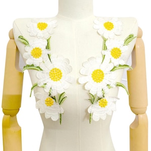 May include: White daisy flower appliques with yellow centers and green leaves are arranged on a cream-colored mannequin. The appliques are embroidered and appear to be made of fabric. The daisies are of varying sizes.