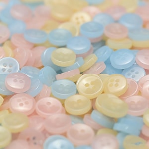 May include: A close-up of a pile of pastel buttons in pink, blue, and yellow. The buttons are circular with four holes and a slightly curved surface. The buttons are scattered, creating a soft, textured appearance.