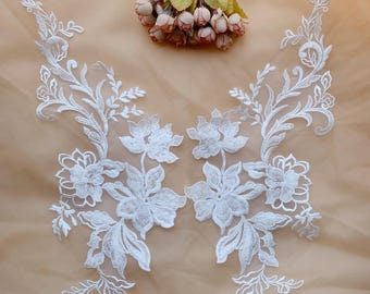 May include: Two white floral lace appliques with intricate designs of flowers and leaves. The appliques are symmetrical and appear to be made of delicate, embroidered fabric. A small bouquet of pink and green flowers is in the background.