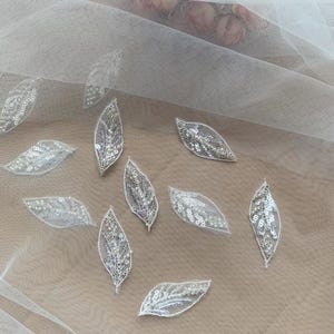 Pode incluir: Close-up de tecido de tule branco com aplicações em forma de folha espalhadas. Cada aplicação apresenta um design delicado e vazado preenchido com lantejoulas prateadas brilhantes. As folhas variam em tamanho e são uniformemente espaçadas.