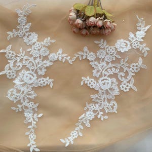 Puede incluir: Dos apliques de encaje floral blanco con intrincados diseños de flores y hojas. Los apliques están dispuestos sobre un fondo beige claro, con un pequeño ramo de flores rosas en la esquina superior derecha.