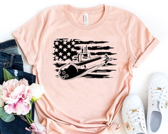 May include: A light pink t-shirt featuring a black and white graphic of a submarine superimposed over a stylized American flag. The shirt has short sleeves and a crew neck. The shirt is displayed with blue jeans, white sneakers, and flowers.