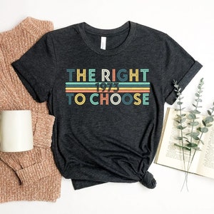 Op de afbeelding: Een donkergrijs T-shirt met de tekst "THE RIGHT 1973 TO CHOOSE" in kleurrijke retro-lettertypen.