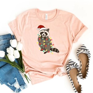 May include: A pink t-shirt with a cartoon raccoon wearing a Santa hat and tangled in Christmas lights.