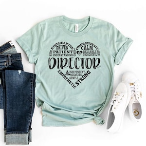 Op de afbeelding: Een lichtblauw T-shirt met een zwart hartvormig ontwerp. Het hart is gevuld met woorden die positieve eigenschappen beschrijven, waaronder "warm", "goedhartig", "meelevend", "gedreven", "geduldig", "begripvol", "kalm", "betrouwbaar", "nadenkend", "trouw", "onafhankelijk", "zelfloos", "vriendelijk", "empatisch" en "sterk". Het woord "DIRECTEUR" staat in grote letters over het hart.