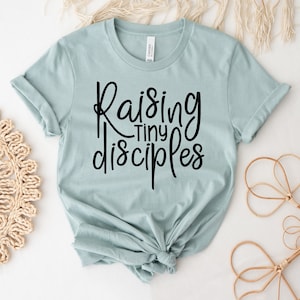 Könnte beinhalten: Ein hellblaues T-Shirt mit dem Text "Raising Tiny Disciples" in schwarzer Schrift.