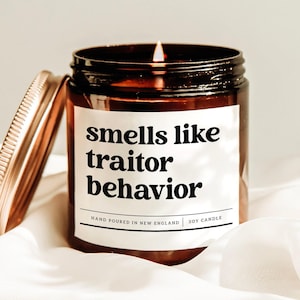 May include: A lit soy candle in a brown glass jar with a copper lid. The label reads "smells like traitor behavior." The candle is hand-poured in New England.