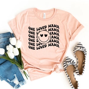 Puede incluir: Una camiseta rosa con el texto "ONE LOVED MAMA" repetido en una fuente retro. Una cara sonriente negra con corazones por ojos está centrada en la camiseta.