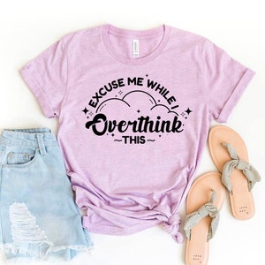 Peut inclure: Un t-shirt rose pâle avec le texte "Excuse me while I overthink this" en lettres noires. Le texte est entouré d'étoiles et de nuages.
