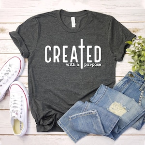 Könnte beinhalten: Ein dunkelgraues T-Shirt mit einem weißen Aufdruck, der "CREATED with a purpose" und ein weißes Kreuz zeigt.