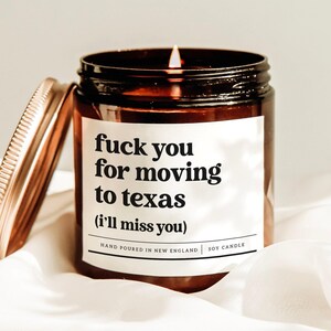 May include: A lit soy candle in a brown glass jar with a gold lid. The label reads "fuck you for moving to texas (i'll miss you)". The candle is hand-poured in New England.