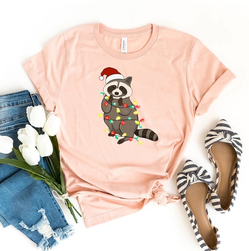 May include: A pink t-shirt with a cartoon raccoon wearing a Santa hat and tangled in Christmas lights.