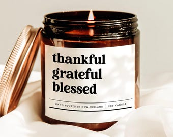 May include: A lit soy candle in a brown glass jar with a gold lid. The label reads "thankful grateful blessed" in black text. The candle is hand-poured in New England.