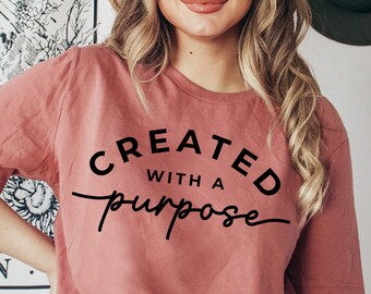 Puede incluir: Una mujer lleva una camiseta rosa polvorienta con el texto "CREATED WITH A purpose" en letras negras. Lleva un sombrero marrón y unos vaqueros azules.