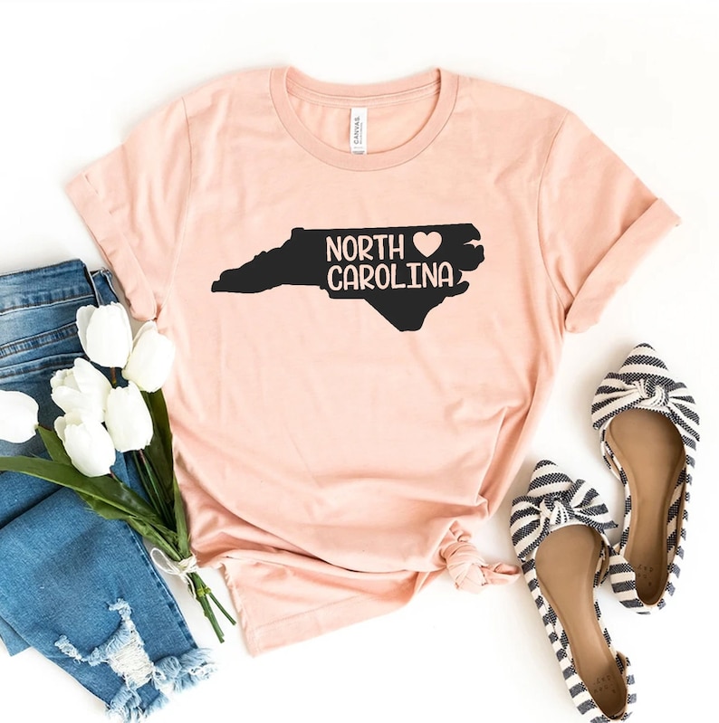 以下が含まれることがあります： ピンクのTシャツに、ノースカロライナ州の黒いアウトラインと「NORTH CAROLINA」の文字が書かれており、その間にハートマークがあります。