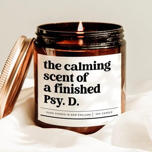 May include: A lit soy candle in a brown glass jar with a gold lid. The label reads "the calming scent of a finished Psy. D." and "Hand Poured in New England". The candle sits on a white cloth.